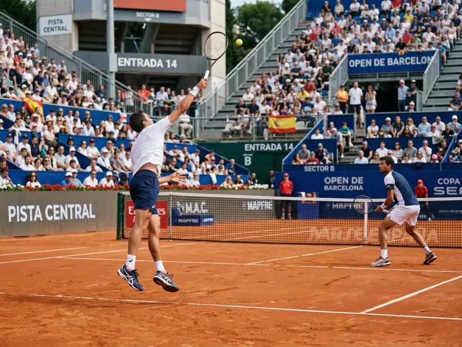 Pista de tenis de tierra batida con dos jugadores en pleno partido, vista lateral desde las gradas